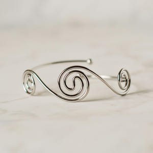 May include: A silver-toned metal cuff bracelet featuring a continuous wire design that forms three distinct spiral shapes. The bracelet has an open-ended construction with a smooth, polished finish and a rounded wire profile throughout the piece.