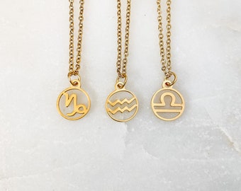 May include: Three gold-colored necklaces with circular pendants featuring zodiac symbols. The pendants are suspended from delicate chains. The symbols represent Capricorn, Aquarius, and Libra. The necklaces are displayed on a white surface.