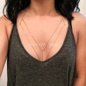 A woman wearing a stylish handmade gold stainless steel chain bra body chain, layered beneath a low-cut gray tank top. The gold body chain features a double layered chain arrangement across the chest. Brand is Sincerely, Jane