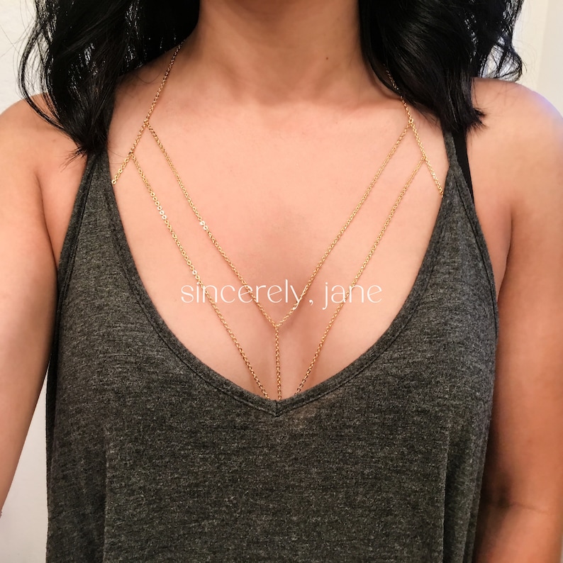 A woman wearing a stylish handmade gold stainless steel chain bra body chain, layered beneath a low-cut gray tank top. The gold body chain features a double layered chain arrangement across the chest. Brand is Sincerely, Jane