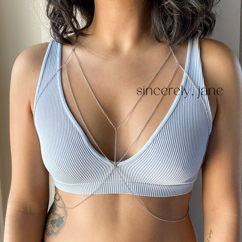 A woman wearing a stylish handmade gold stainless steel chain bra body chain, layered on top of a a low-cut light blue bralette. The silver body chain features a double layered chain arrangement across the chest. Brand is Sincerely, Jane
