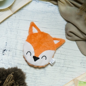 Könnte beinhalten: Ein orangefarbener Plüschfuchs-Kopf mit weißem Gesicht und schwarzen Augen. Der Fuchs hat einen fröhlichen Ausdruck und liegt auf einer weißen Holzoberfläche.