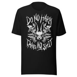 以下が含まれることがあります： 黒いTシャツに白いグラフィック。デザインは、矢印を持つウサギと、かすれたフォントで「DO NO HARM TAKE NO SHIT」というテキストが特徴です。グラフィックはTシャツの前面の中央に配置されています。