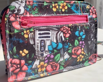 Può includere: Un portafoglio nero con cerniera con una stampa floreale colorata che presenta un personaggio di Star Wars, R2-D2, e una cerniera rosa.