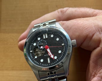 May include: A stainless steel wristwatch with a black face and a silver band. The watch has white hour markers, a red second hand, and a small window revealing the inner workings. The word "AUTOMATIC" is visible on the watch face.