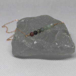 Puede incluir: Un collar de cadena dorada con una fila de siete cuentas de piedras preciosas multicolores. Las cuentas son una mezcla de verde, marrón, rosa y blanco.