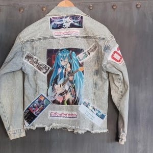 Op de afbeelding: Een lichtblauw denim jack met verschillende patches, waaronder een groot beeld van een anime-personage met blauw haar dat gitaar speelt, en de tekst "Miku Hatsune".