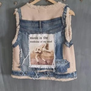 Op de afbeelding: Een spijkerbroekvest met een beige imitatiebontvoering en -afwerking. Het vest heeft een patch met de tekst "music is the medicine of my mind" en een afbeelding van een persoon die gitaar speelt. Het vest hangt aan een houten hanger.
