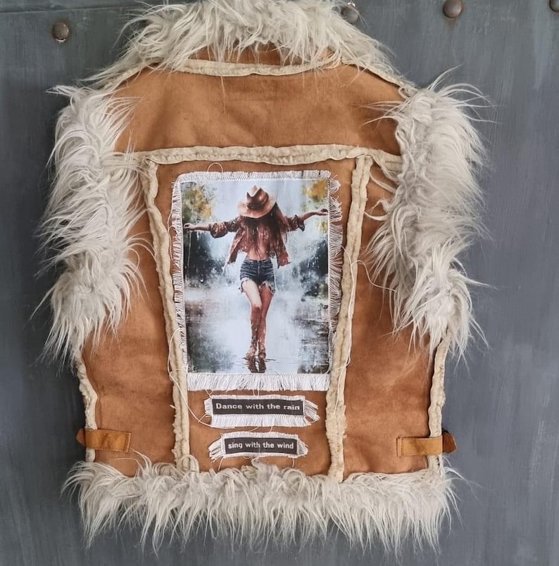 May include: Brown suede vest with cream faux fur trim. The back has a framed image of a person in a hat, arms outstretched in the rain. Text on the vest reads "Dance with the rain" and "sing with the wind."