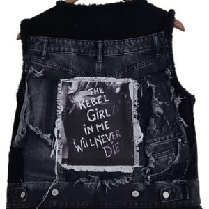 Op de afbeelding: Zwarte denim vest met een distressed design en een zwarte imitatiebont kraag. De vest heeft een patch met de tekst "THE REBEL GIRL IN ME WILL NEVER DIE" in witte letters. De vest hangt aan een houten hanger.