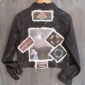 May include: A dark grey denim jacket adorned with patches and embellishments. Patches feature the text "HARRY STYLES", "WE BELONG TOGETHER", "TOUR 2026", and "TIME, KISS ALL, DISCO, OCCASIONALLY". The jacket is embellished with small rhinestones.
