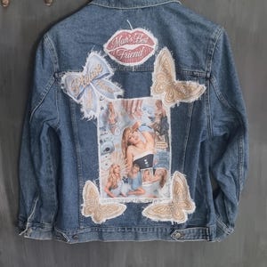 Op de afbeelding: Een blauw spijkerjack met een grote centrale collage van Sabrina Carpenter-afbeeldingen, omringd door patches met gerafelde randen, waaronder een roze lipvorm met de tekst Man's Best Friend, een lichtblauwe strik en vier beige vlinderapplicaties.