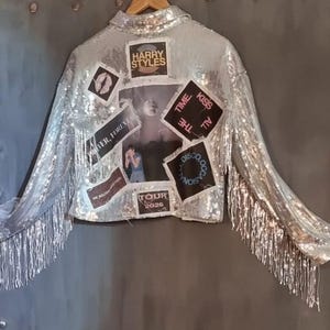 May include: Silver sequined jacket with fringed sleeves. Patches include "Harry Styles", "Time Kiss All The", "Disco Occasion", "Tour 2026", and other graphics. The jacket is displayed on a wooden hanger.