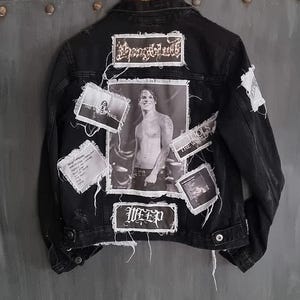 Op de afbeelding: Zwarte spijkerjas met meerdere patches en een centrale zwart-wit afbeelding. Patches bevatten tekst en een foto van een persoon. De jas heeft een knoopsluiting en distressed details.