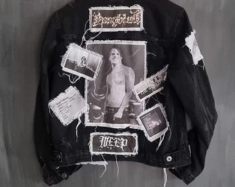 May include: Black denim jacket adorned with patches and a central black and white image. Patches feature text and a photograph of a person. The jacket has a button closure and distressed detailing.