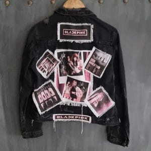 Op de afbeelding: Zwarte spijkerjas met meerdere patches met de bandnaam "BLACKPINK", foto's en tourdetails. De jas heeft een versleten look met gerafelde randen en een knoopsluiting.