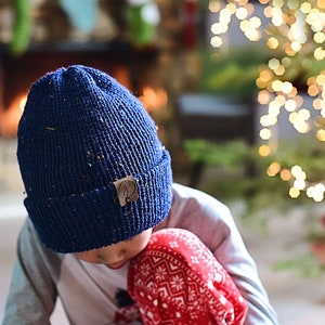 Può includere: Un bambino indossa un berretto blu a maglia con una toppa in pelle marrone e un maglione a motivi rossi e bianchi è seduto davanti a un albero di Natale con le luci.