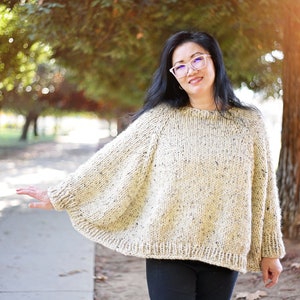 May include: A woman wearing a cream-colored knitted sweater with a speckled pattern. The sweater has a wide, flowing silhouette and is worn with black pants.
