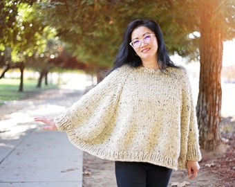 May include: A woman wearing a cream-colored knitted sweater with a speckled pattern. The sweater has a wide, flowing silhouette and is worn with black pants.