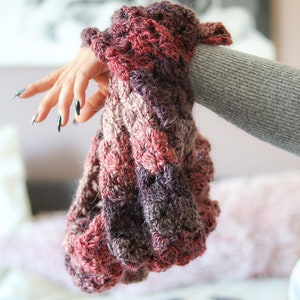 May include: A close-up of a hand holding a crocheted scarf with a pink, purple and brown colour gradient.