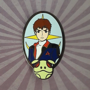 Könnte beinhalten: Eine blaue und rote Emaille-Anstecknadel mit einer Comicfigur mit braunen Haaren und einer blauen Jacke. Die Figur hält einen gelben und weißen Roboter vor sich. Die Anstecknadel ist vor einem weißen Hintergrund mit einem gelben Stern platziert.