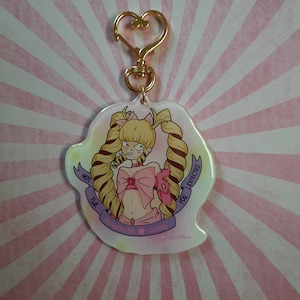 Könnte beinhalten: Ein rosa-weißer Charakter-Anhänger mit einem goldenen Schlüsselanhänger. Der Charakter hat blonde Haare, eine rosa Schleife und ein rosa-blaues Outfit. Der Text auf dem Anhänger lautet "Nicht das magische Mädchen, das du erwartet hast."