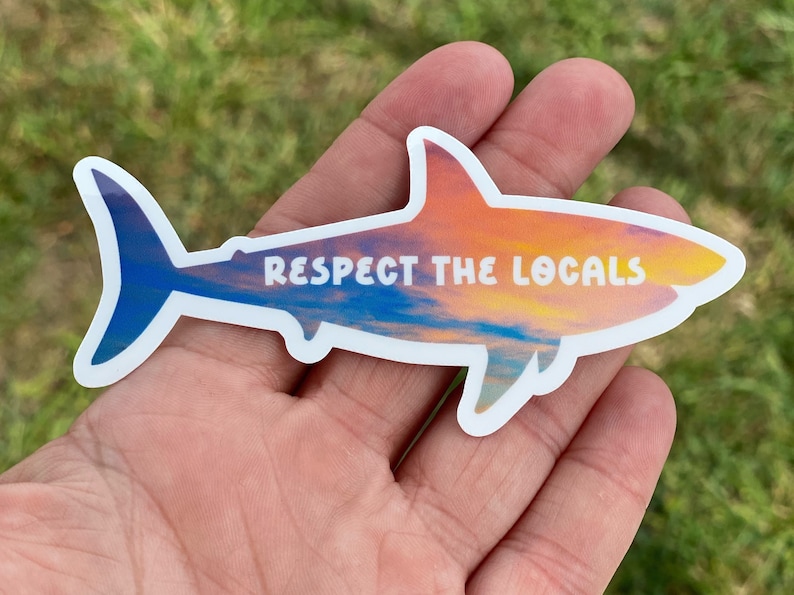 May include: A white shark silhouette sticker with a sunset sky background. The sticker says "Respect the Locals" in white text.