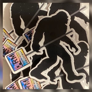 May include: A stack of stickers featuring a black silhouette of Bigfoot walking with a stick. The stickers have a rainbow gradient background with the text "Robert Moses Causeway" and "RM" in white.