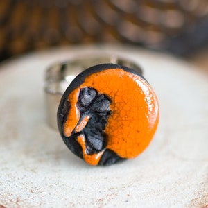 Peut inclure: Une bague en céramique noire et orange avec un glaçage craquelé. La bague est posée sur une surface blanche.