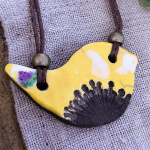 Peut inclure: Un pendentif en céramique jaune en forme d'oiseau avec des détails noirs et blancs suspendu à un collier en cordon marron. Le pendentif est sur une surface en tissu gris clair.