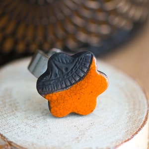 Può includere: Un anello in ceramica nero e arancione a forma di stella. L'anello è su una superficie di legno.