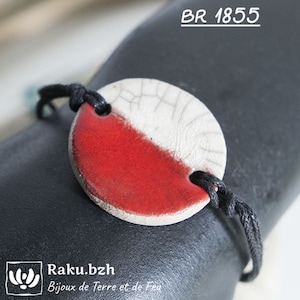 Peut inclure: Un bracelet en cordon noir avec un pendentif rond en céramique rouge et blanche. Le pendentif est recouvert d'un glaçage blanc craquelé sur une moitié et d'un glaçage rouge uni sur l'autre moitié. Le bracelet est noué et porte le texte "BR 1855" et "Raku.bzh Bijoux de Terre et de Feu" sur le fond.
