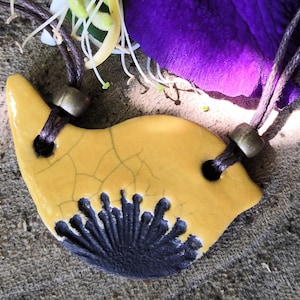 Peut inclure: Un pendentif en céramique jaune avec un motif noir, suspendu à un cordon brun avec des perles métalliques. Le pendentif a un glaçage craquelé.
