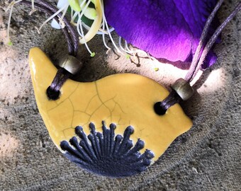 Può includere: Un ciondolo in ceramica giallo con un design nero, appeso a un cordoncino marrone con perline metalliche. Il ciondolo ha una smaltatura craquelata.