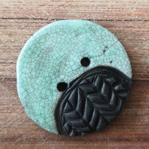 Può includere: Un bottone rotondo in ceramica con una glassa verde chiaro e un design nero testurizzato. Il bottone ha due fori per cucire.