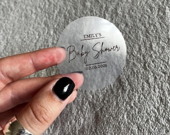 May include: A clear round sticker with black text that says "Emily's Baby Shower 02.05.2025".