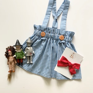 May include: A blue and white gingham overall skirt with brown buttons and a red polka dot bow. The skirt is laying on a white surface with three small dolls.
