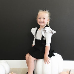 May include: A young girl wearing a black overall skirt with ruffle sleeves sits on a large white pumpkin. She is smiling and looking at the camera. There are several smaller pumpkins in the background.