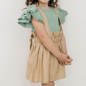 May include: A young girl wearing a light green ruffled top with a brown skirt and a white lace bow in her hair.