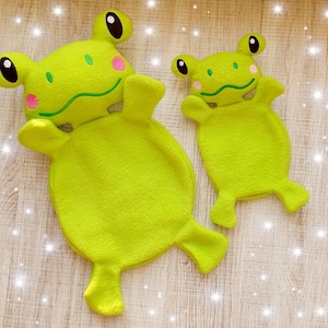 May include: Two green frog-shaped plush toys. The larger frog is lying down with its legs outstretched. The smaller frog is sitting up with its legs crossed. Both frogs have embroidered eyes and mouths.
