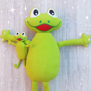 May include: Two green plush frog toys. The larger frog has a red mouth and is holding a smaller frog with its arms.
