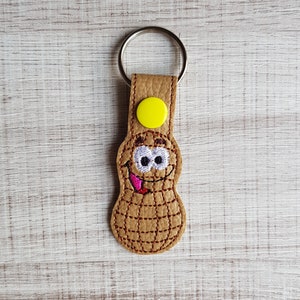 May include: A brown leather peanut-shaped keychain with a smiling face, big eyes, and a pink tongue sticking out. The keychain has a yellow button and a silver key ring.