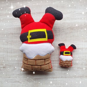 May include: Two Christmas decorations in the shape of Santa Claus. The larger one measures approximately 25 cm tall, and the smaller one is about 10 cm. Both feature red legs with black boots, a black belt with a gold buckle, and a white chimney with a brown brick base.