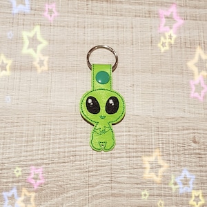 May include: Green alien keychain with big eyes and a friendly smile. The keychain is made of felt and has a silver metal ring.