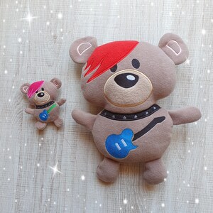 May include: Two plush teddy bears, one large and one small. The larger bear is brown with a red mohawk and a blue guitar embroidered on its chest. The smaller bear is brown with pink ears and a blue guitar embroidered on its chest.