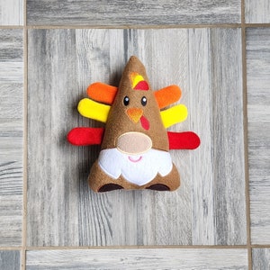 May include: A plush toy shaped like a gnome with a turkey-themed design. The gnome has a brown felt body, a white beard, and a red, yellow, and orange feathered headdress.