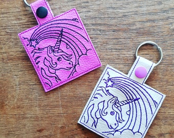 May include: Two keychains featuring a unicorn and rainbow design. One keychain is pink with a black unicorn and rainbow, the other is white with a purple unicorn and rainbow.