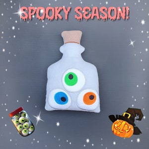 Könnte beinhalten: Weiße Filz-Flasche mit drei Filz-Augen, einem grünen, einem blauen und einem orangefarbenen. Die Flasche ist mit "Spooky Season!" beschriftet und von einem weißen Hintergrund mit silbernem Glitzer umgeben.