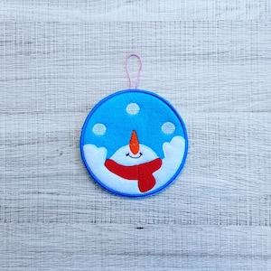 May include: A blue felt ornament with a snowman design. The snowman has a red scarf, a carrot nose, and two black eyes. The ornament has a pink string for hanging.