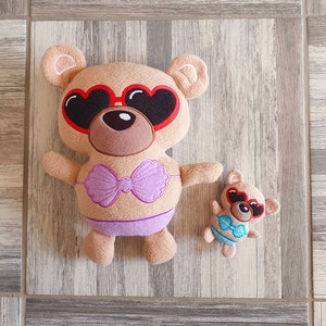Può includere: Due orsacchiotti di peluche marroni che indossano occhiali da sole e costumi da bagno. L'orso più grande indossa un costume da bagno viola e l'orso più piccolo indossa un costume da bagno blu.
